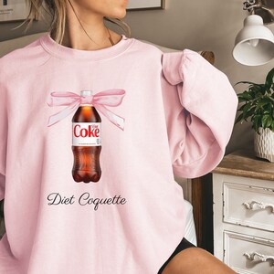 Puede incluir: Sudadera rosa con un gr&aacute;fico de una botella de Coca-Cola Light con un lazo rosa y el texto "Diet Coquette".