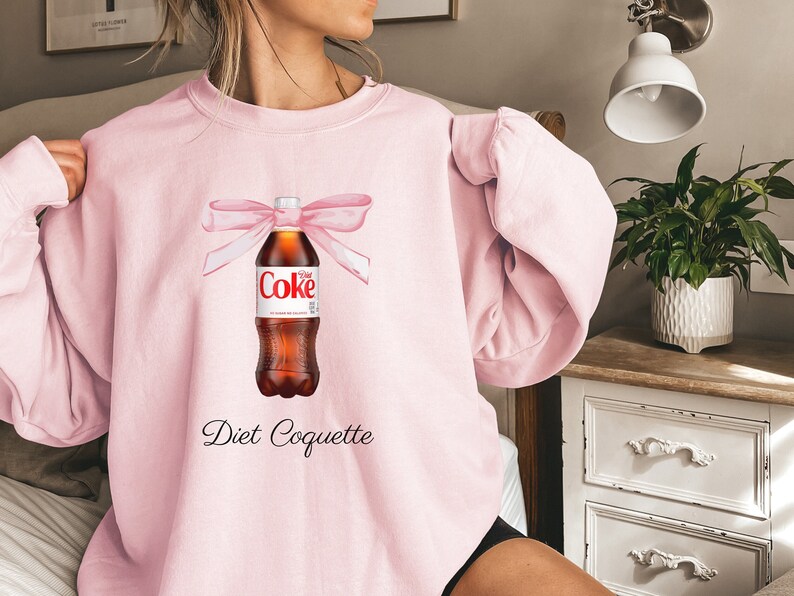 Puede incluir: Sudadera rosa con un gr&aacute;fico de una botella de Coca-Cola Light con un lazo rosa y el texto "Diet Coquette".