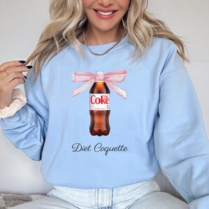 Puede incluir: Una sudadera de color azul claro con un gr&aacute;fico de una botella de Coca-Cola Light atada con una cinta rosa. El texto "Diet Coquette" est&aacute; impreso debajo de la botella.
