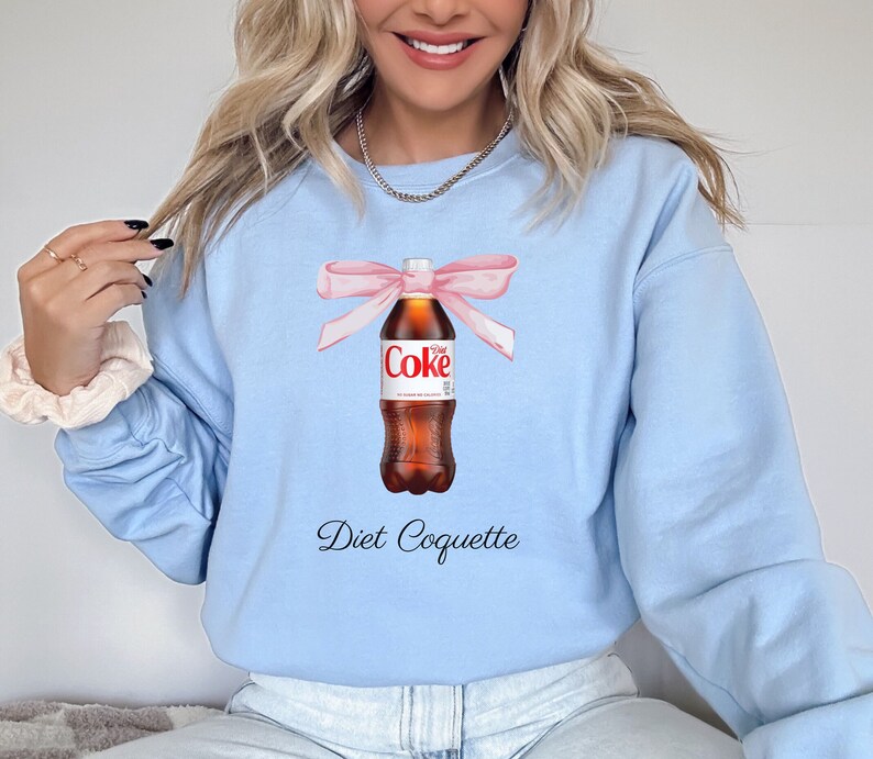 Puede incluir: Una sudadera de color azul claro con un gr&aacute;fico de una botella de Coca-Cola Light atada con una cinta rosa. El texto "Diet Coquette" est&aacute; impreso debajo de la botella.