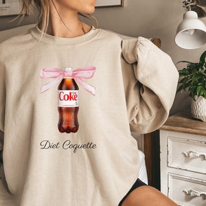 Puede incluir: Una sudadera beige con un lazo rosa atado alrededor de una botella de Coca-Cola Light. El texto "Diet Coquette" est&aacute; impreso debajo de la botella.