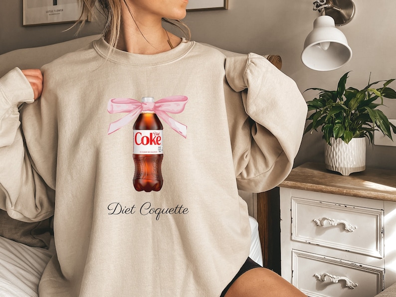 Puede incluir: Una sudadera beige con un lazo rosa atado alrededor de una botella de Coca-Cola Light. El texto "Diet Coquette" est&aacute; impreso debajo de la botella.