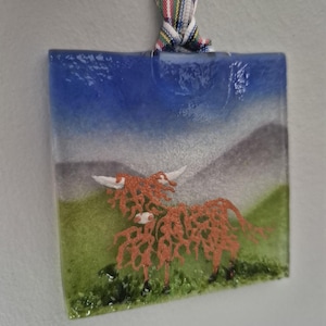 May include: A glass ornament featuring a cartoon Highland cow with a blue sky, green grass, and white mountains in the background.