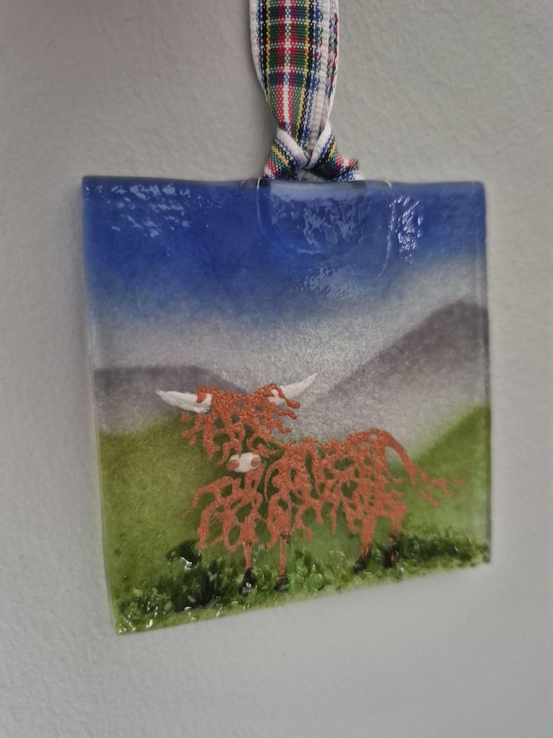 May include: A glass ornament featuring a cartoon Highland cow with a blue sky, green grass, and white mountains in the background.