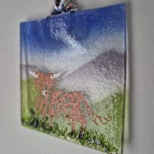 May include: A glass bauble depicting a pink Highland cow standing in a green field with a blue sky and white clouds in the background.