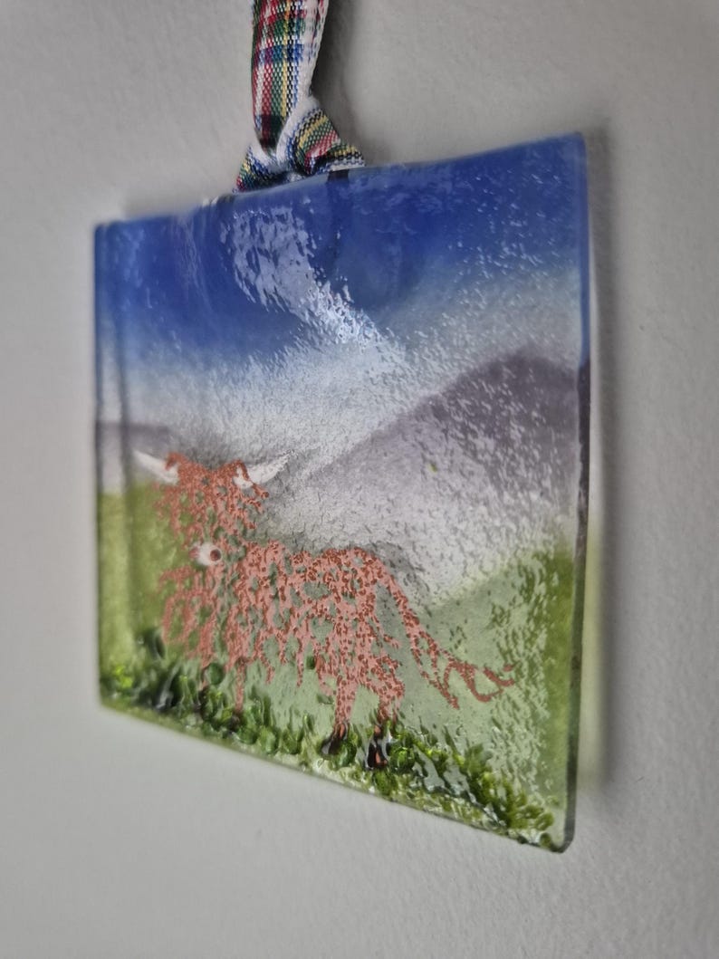 May include: A glass bauble depicting a pink Highland cow standing in a green field with a blue sky and white clouds in the background.