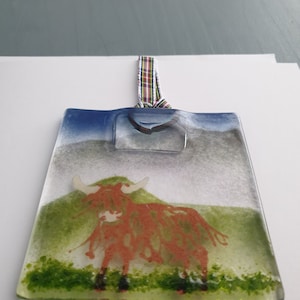 May include: A glass ornament featuring a red Highland cow standing in a green field with a blue sky in the background. The ornament has a tartan ribbon attached to the top.