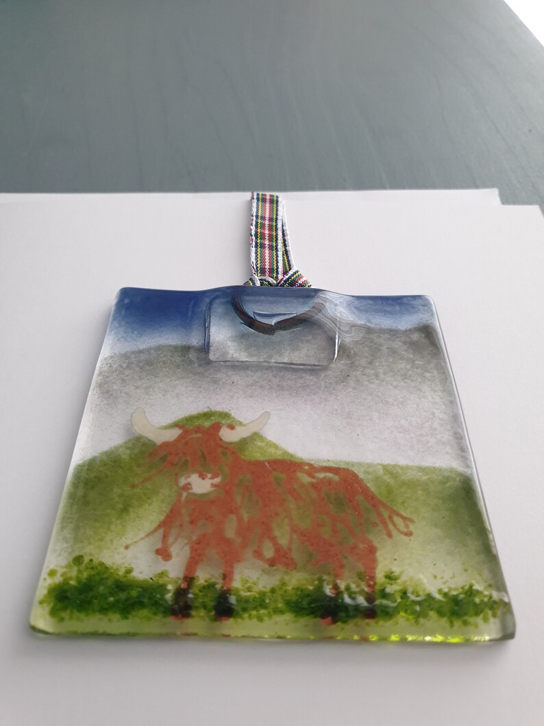 May include: A glass ornament featuring a red Highland cow standing in a green field with a blue sky in the background. The ornament has a tartan ribbon attached to the top.