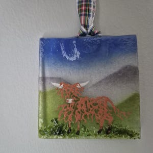 May include: A glass bauble depicting a red Highland cow against a backdrop of a blue sky and green grass. The bauble is suspended from a red, white and green tartan ribbon.