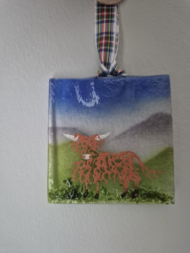 May include: A glass bauble depicting a red Highland cow against a backdrop of a blue sky and green grass. The bauble is suspended from a red, white and green tartan ribbon.