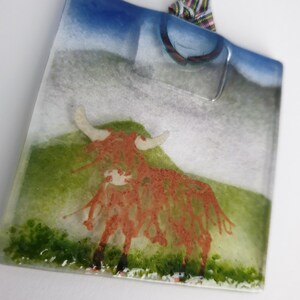 May include: A square glass ornament featuring a red Highland cow standing in a green field. The ornament has a blue sky and white clouds in the background. It is hanging from a red and white checked ribbon.