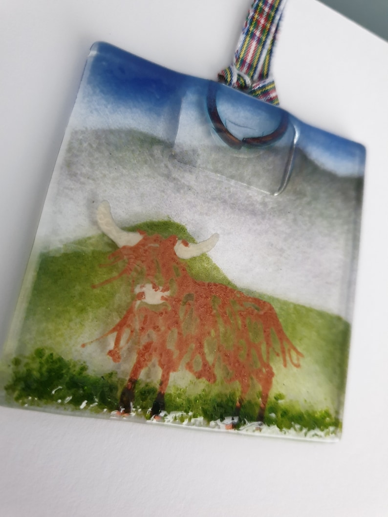 May include: A square glass ornament featuring a red Highland cow standing in a green field. The ornament has a blue sky and white clouds in the background. It is hanging from a red and white checked ribbon.