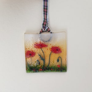 May include: A square glass ornament with a poppy flower design. The ornament has a yellow and white background with red poppies and green foliage. A plaid ribbon is attached for hanging.