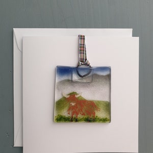 May include: A glass bauble depicting a red Highland cow standing in a green field with a blue sky and white clouds in the background. The bauble is attached to a red, green and white tartan ribbon.