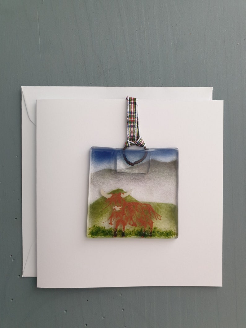 May include: A glass bauble depicting a red Highland cow standing in a green field with a blue sky and white clouds in the background. The bauble is attached to a red, green and white tartan ribbon.