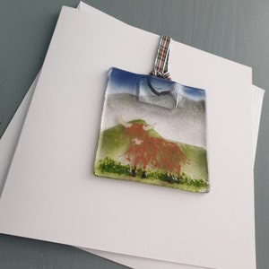 May include: A glass bauble depicting a highland cow with a reddish-brown body and white horns, standing on a green field against a blue sky. The bauble is attached to a white ribbon with a tartan pattern.