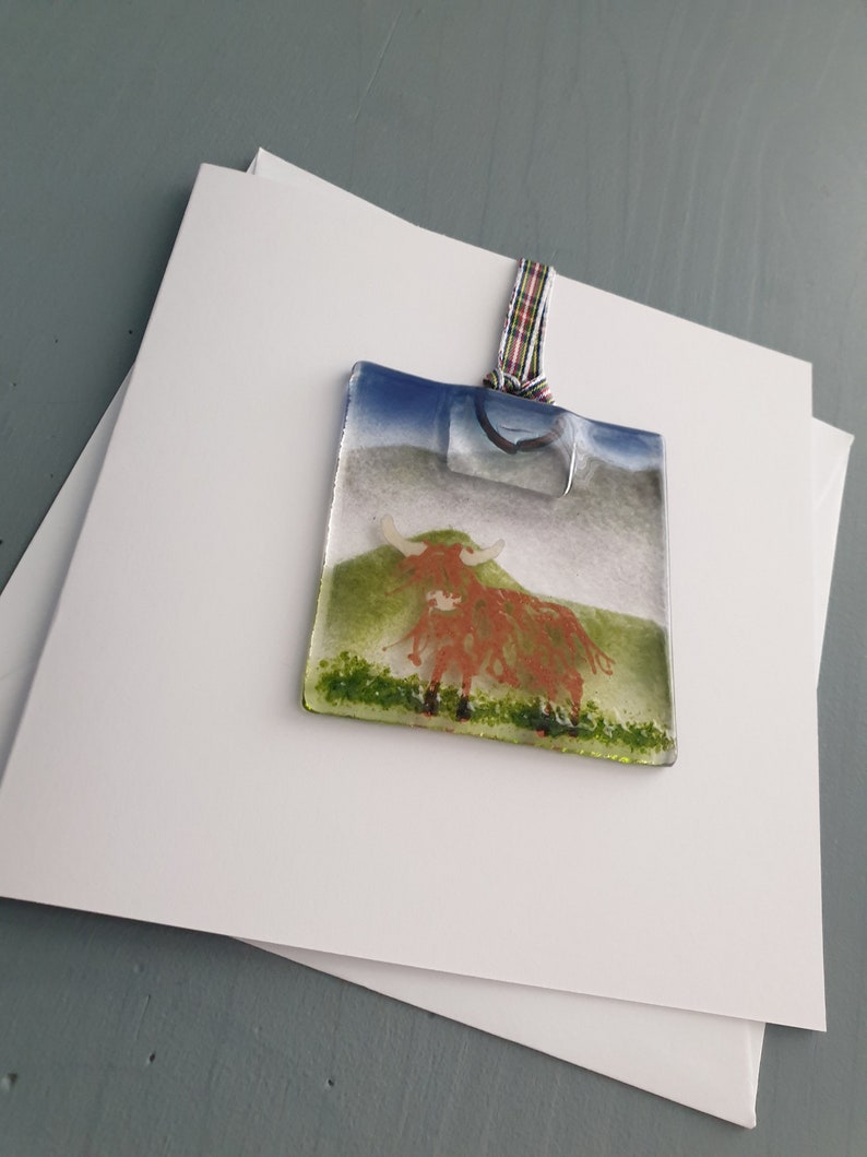 May include: A glass bauble depicting a highland cow with a reddish-brown body and white horns, standing on a green field against a blue sky. The bauble is attached to a white ribbon with a tartan pattern.
