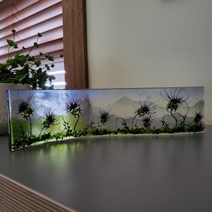 May include: A curved glass art piece depicting a landscape with stylized purple thistle flowers and green foliage. The background shows a mountain range under a blue and white sky. The piece is approximately 30 cm long.