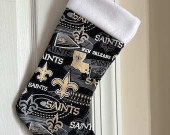 May include: A black and gold Christmas stocking with the text "SAINTS" and "NEW ORLEANS" printed on it. The stocking has a white cuff and is hanging on a white door.