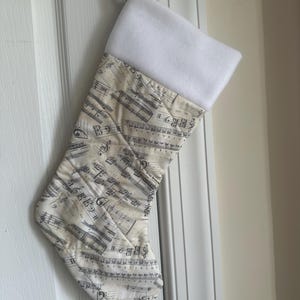 May include: A Christmas stocking with a white cuff and a body patterned with musical notes. The stocking is hanging on a white door. The fabric has a cream background with black musical notation.