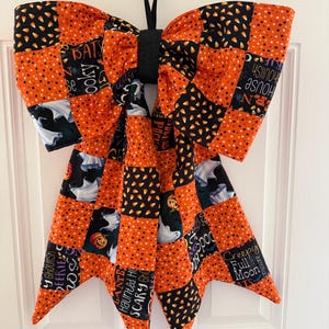 Könnte beinhalten: Eine große dekorative Schleife aus Stoff mit einem Patchwork-Muster aus Halloween-Motiven wie Geistern, Kürbissen, Zuckermais und Texten wie Spooky, Bats, Creepy Full Moon und Haunted House in den Farben Orange, Schwarz und Weiß.