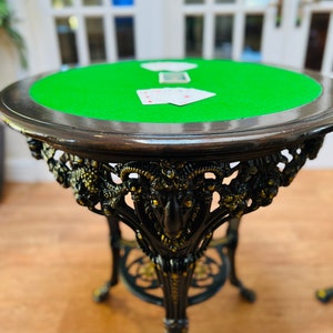 May include: A black cast iron table with a green felt top. The table features a decorative ram's head design and ornate legs. There are three playing cards on the table.