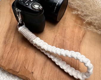 Può includere: Fotocamera digitale Fujifilm nera con cinturino da polso in macramè color crema. La fotocamera è su una tavola di legno. Il cinturino da polso è in cotone intrecciato e ha una chiusura argentata.