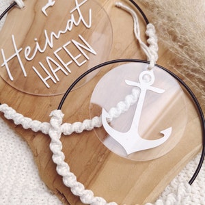 Op de afbeelding: Twee transparante acryl cirkels met witte letters en een wit ankerontwerp. De cirkels hangen aan een wit touw op een houten oppervlak.