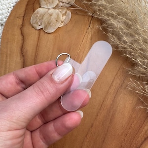 Könnte beinhalten: Ein klarer, rechteckiger Handyhalter mit einem silbernen Ring. Der Halter wird vor einem Holzuntergrund gehalten, mit getrockneten Blumen und einer weißen Strickdecke im Hintergrund. Der Halter ist für die Befestigung an der Rückseite eines Telefons konzipiert.
