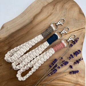 May include: Three keychains with white rope and different colored accents. The keychains are on a wooden surface with lavender flowers.