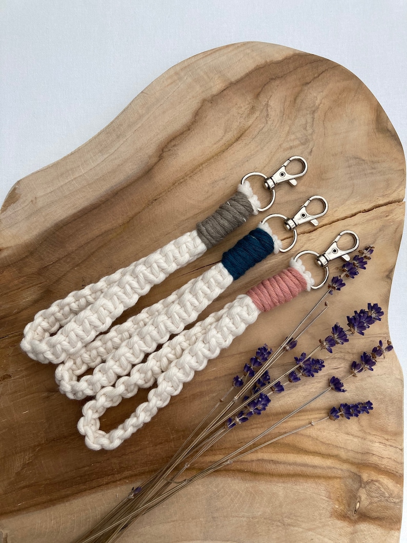 May include: Three keychains with white rope and different colored accents. The keychains are on a wooden surface with lavender flowers.