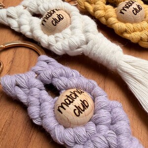 May include: A selection of macrame keyrings in white, yellow, lavender, and teal. Each keyring has a wooden bead with the words "Matcha Club" burned into it. The keyrings are finished with gold-coloured metal clasps and are displayed on a wooden surface.