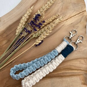 May include: A white and light blue macrame keychain with a dark blue accent and silver key rings. The keychain is on a wooden surface with dried wheat and lavender.