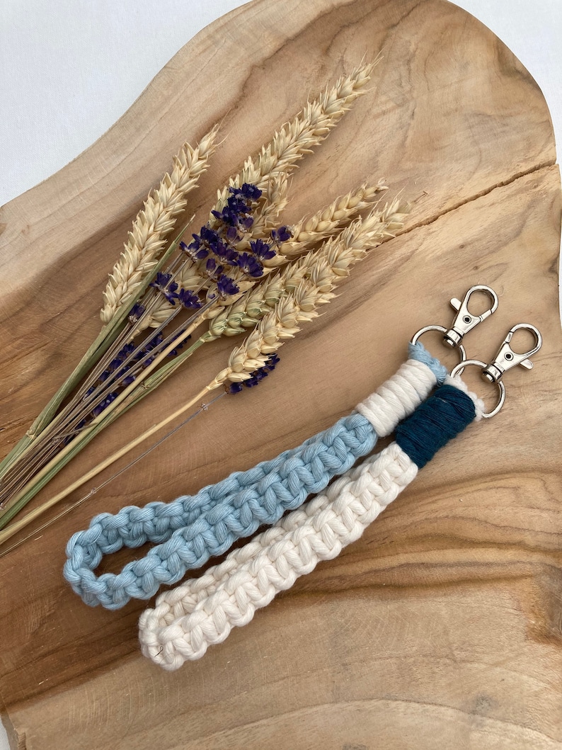 May include: A white and light blue macrame keychain with a dark blue accent and silver key rings. The keychain is on a wooden surface with dried wheat and lavender.
