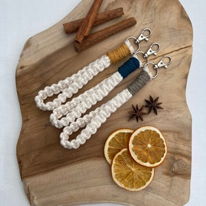 May include: Three white macrame keychains with different colored accents. The keychains are on a wooden surface with cinnamon sticks and dried orange slices.