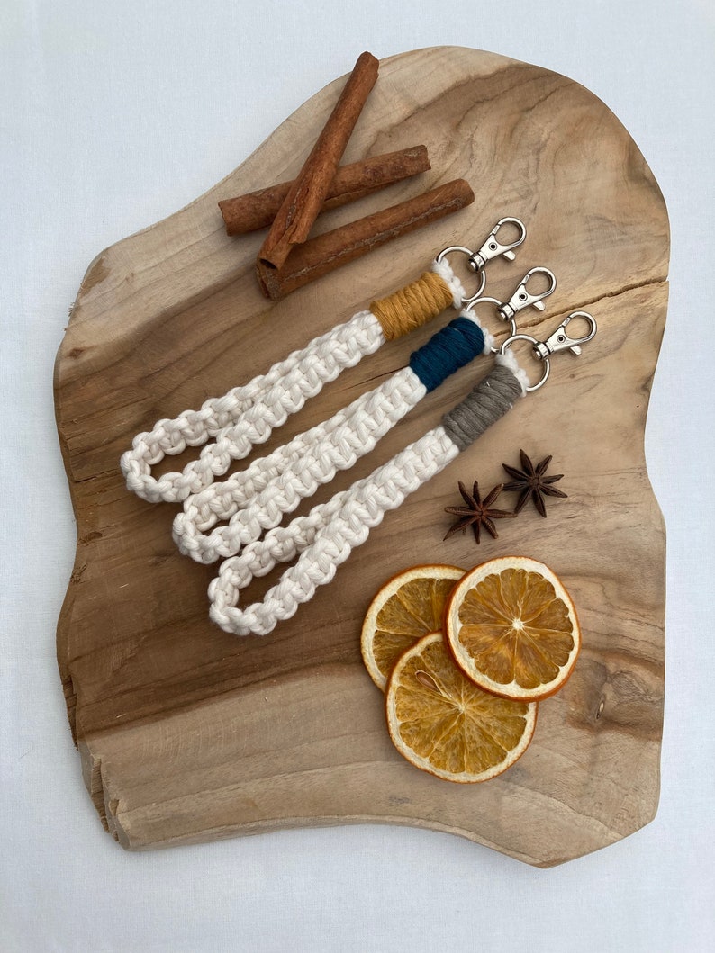 May include: Three white macrame keychains with different colored accents. The keychains are on a wooden surface with cinnamon sticks and dried orange slices.