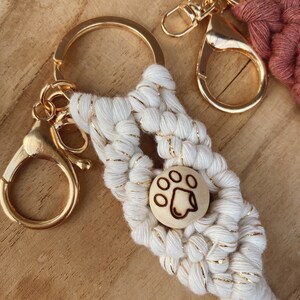Op de afbeelding: Witte en gouden macramé sleutelhanger met een houten hondenpoot charme. De sleutelhanger heeft een gouden metalen ring en sluiting.