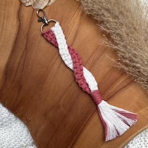 Op de afbeelding: Een macramé sleutelhanger met een gedraaid ontwerp in wit en stoffig roze. De sleutelhanger heeft een zilveren sluiting en een kwastje aan het uiteinde. Het item wordt gepresenteerd op een houten oppervlak.