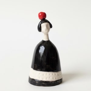 Könnte beinhalten: Keramikfigur einer Person in einem schwarzen Kleid mit weißem Band am unteren Rand. Ein kleiner roter Apfel ruht auf dem Kopf. Die Figur hat ein minimalistisches Design und eine glatte, glänzende Oberfläche.