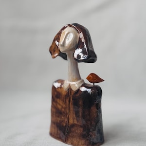 Pode incluir: Uma escultura em cerâmica de uma figura com vestido e cabelo castanho, rosto pálido e um pequeno pássaro laranja pousado no ombro. A figura tem um design minimalista e um acabamento liso e brilhante. A escultura está sobre um fundo neutro.