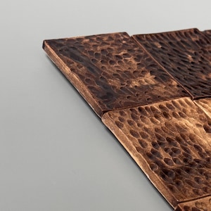 May include: A collection of square tiles featuring a dark brown, metallic finish with a hammered, dimpled texture across the surface. The tiles are arranged in a grid pattern, showcasing a reflective sheen and a rugged, uneven appearance.
