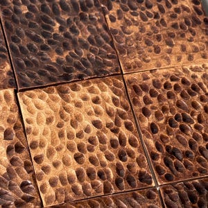 May include: A collection of square metal tiles featuring a hammered, dimpled texture. The surface of each tile displays a warm, metallic copper finish with varying shades of dark brown and bronze highlights across the indented, textured pattern.