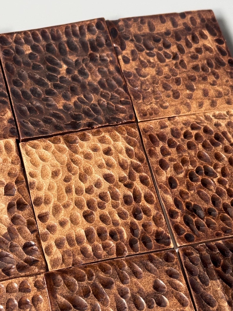 May include: A collection of square metal tiles featuring a hammered, dimpled texture. The surface of each tile displays a warm, metallic copper finish with varying shades of dark brown and bronze highlights across the indented, textured pattern.