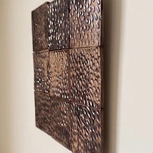 May include: A square wall art piece composed of nine individual square tiles arranged in a three-by-three grid. Each tile features a dark bronze metallic finish with a deeply hammered, dimpled texture that creates a reflective, uneven surface.