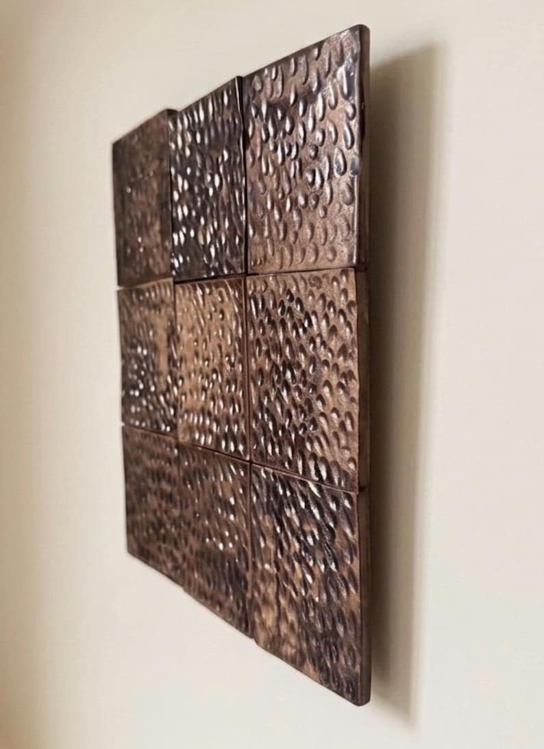 May include: A square wall art piece composed of nine individual square tiles arranged in a three-by-three grid. Each tile features a dark bronze metallic finish with a deeply hammered, dimpled texture that creates a reflective, uneven surface.