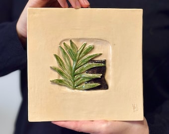 May include: A beige ceramic tile with a raised green leaf design and a black and brown background. The tile is framed by a slightly raised border.