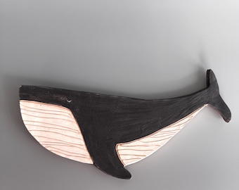 May include: A flat, whale-shaped wall decoration featuring a black upper body and a light pink underbelly with horizontal grooved lines. The whale is depicted in a horizontal swimming position with a prominent tail fluke and a single side fin.