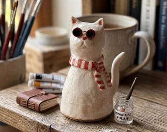 May include: A ceramic cat figurine wearing sunglasses and a red and white striped scarf. The cat is sitting on a wooden surface with a small leather-bound book, paintbrushes, and a jar labeled "MINIATURE POLISH".
