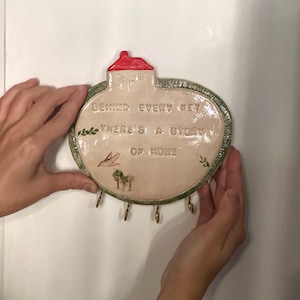 Puede incluir: Un llavero de cerámica con una parte superior en forma de casa y una base circular. El llavero es de color beige con un techo rojo y ribete verde. Tiene el texto "BEHIND EVERY KEY THERE'S A STORY OF HOME" y varios ganchos para llaves.
