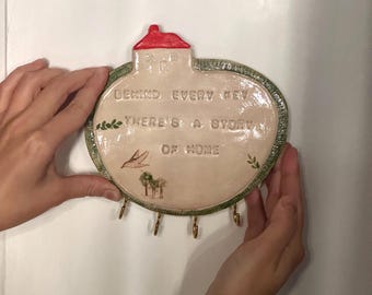 Puede incluir: Un llavero de cerámica con una parte superior en forma de casa y una base circular. El llavero es de color beige con un techo rojo y ribete verde. Tiene el texto "BEHIND EVERY KEY THERE'S A STORY OF HOME" y varios ganchos para llaves.